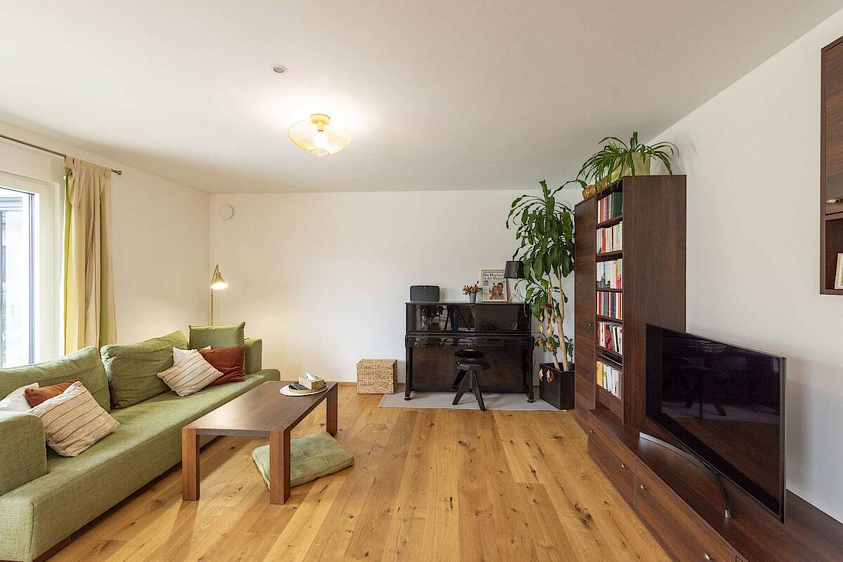 Ein gemütliches Wohnzimmer mit grünem Ecksofa, Couchtisch aus Holz und schwarzem Klavier. Pflanzen und ein Bücherregal runden die wohnliche Atmosphäre ab.