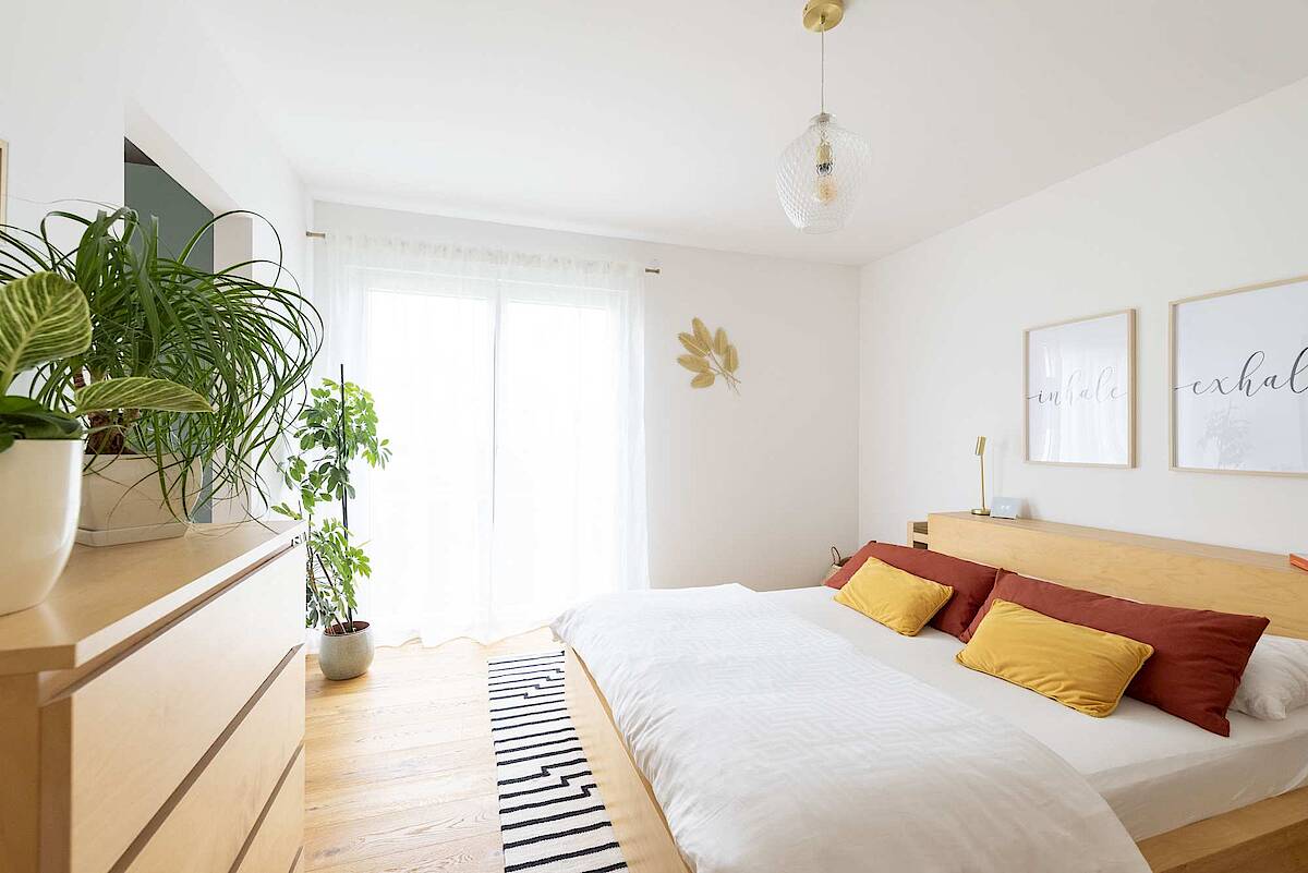 Ein modernes Schlafzimmer mit einem Holz-Doppelbett, senfgelben und terrakottafarbenen Kissen. Pflanzen und helle Vorhänge ergänzen die minimalistische Einrichtung.