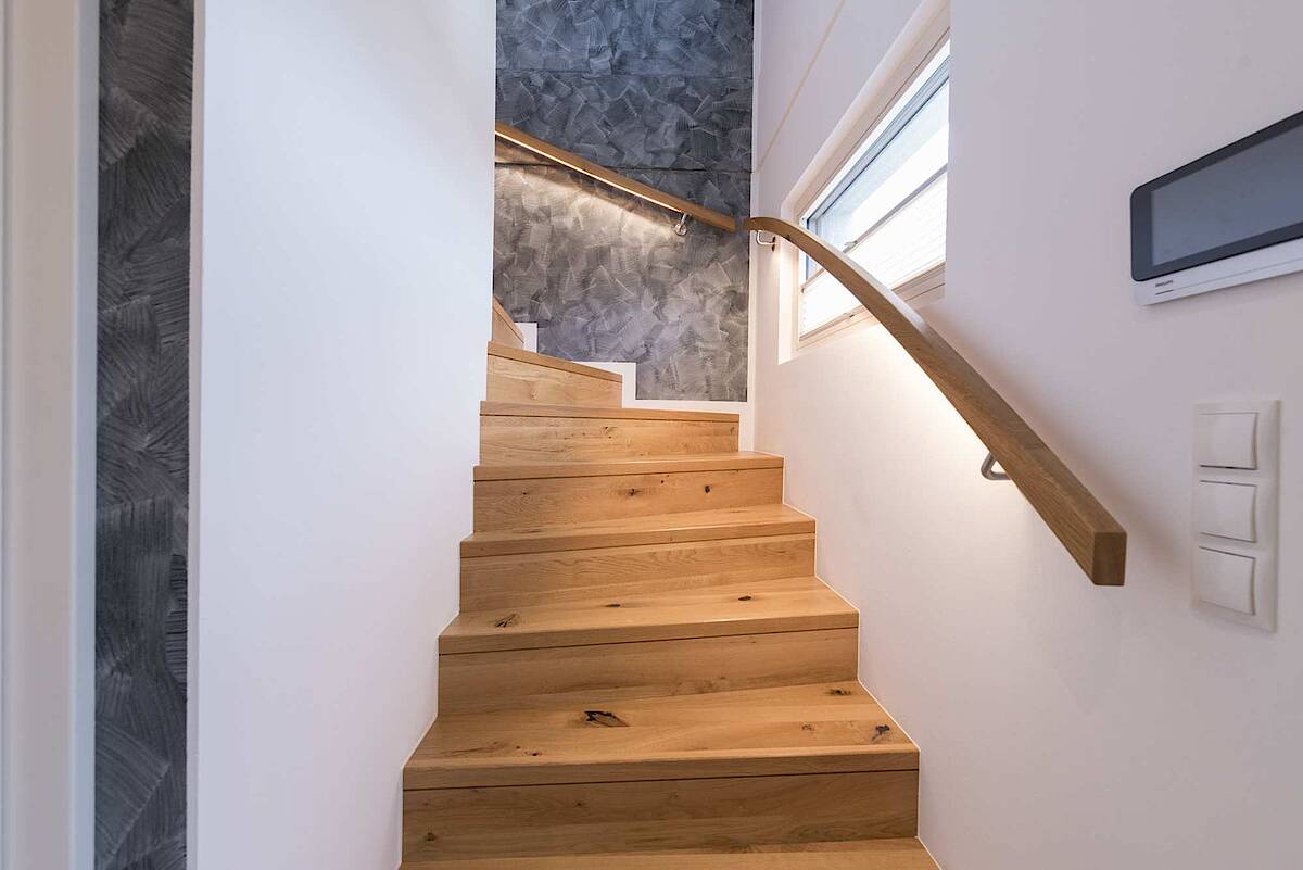 Eine moderne Holztreppe mit einem eleganten Handlauf aus Holz und integrierter Beleuchtung. Die graue Akzentwand im Hintergrund sorgt für einen stilvollen Kontrast.