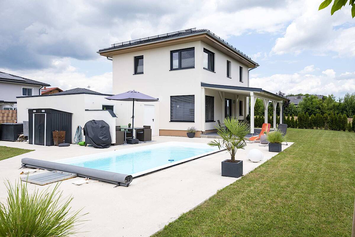 Ein luxuriöser Garten mit einem eleganten Swimmingpool, Sonnenliegen und einem überdachten Sitzbereich. Das moderne Haus und die umliegende Begrünung schaffen eine entspannte Atmosphäre.