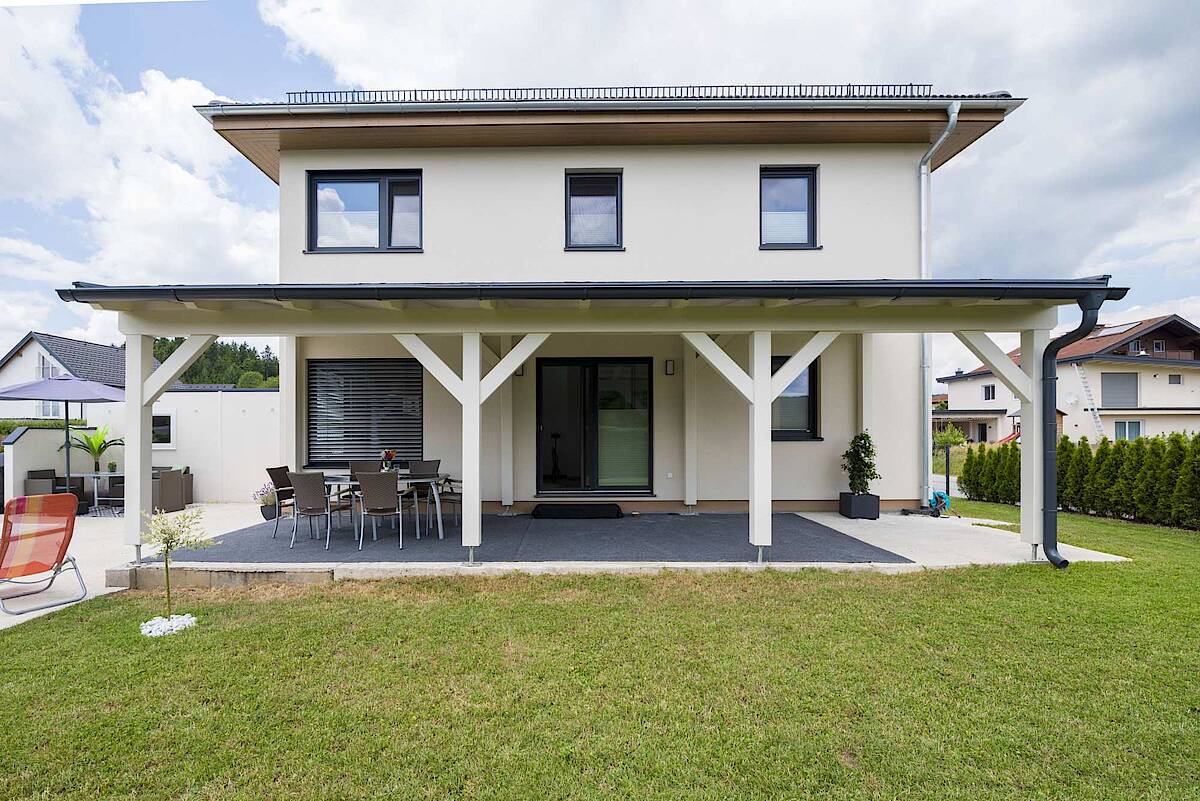 Rückansicht eines modernen Hauses mit einer überdachten Terrasse und Sitzmöglichkeiten im Freien, umgeben von einem großzügigen Rasen und angrenzenden Nachbarhäusern.