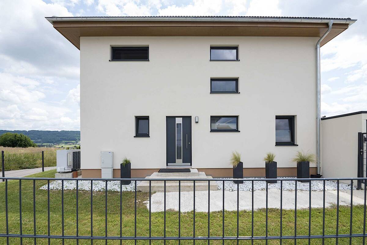 Ein minimalistisches zweistöckiges Haus mit weißer Fassade und modernen Fenstern, umgeben von einem gepflegten Rasen und einem schwarzen Metallzaun.