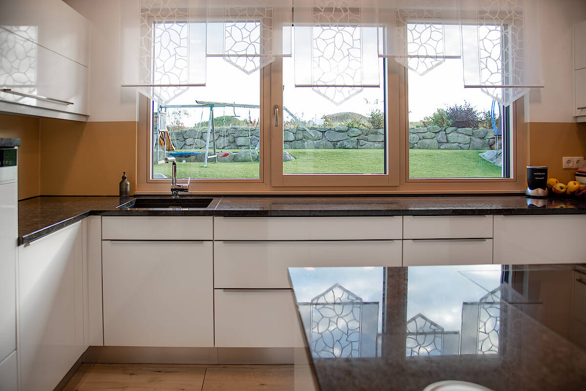 Küche mit Gartenblick: Moderne Küche mit weißem Interieur und schwarzer Arbeitsplatte. Große Fenster bieten Blick auf den grünen Garten mit einer Schaukel.