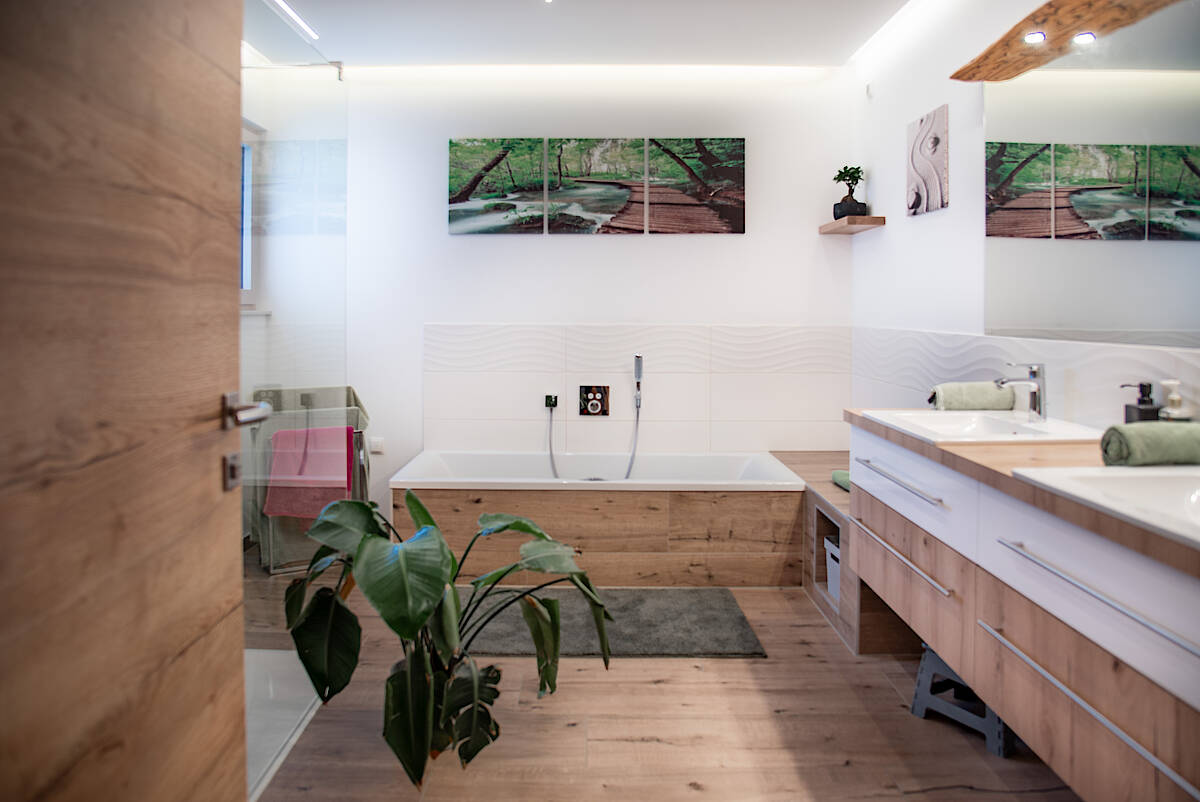 Komplettansicht Badezimmer: Elegantes Badezimmer mit Holzoptik, eingebauter Badewanne, Dusche und grüner Pflanze im Vordergrund. Wanddekoration mit Naturmotiven.
