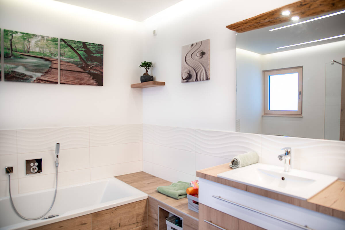 Bad mit Holzverkleidung: Modernes Badezimmer mit eingebauter Badewanne, weißen Fliesen, Holzverkleidung und minimalistischer Dekoration. An der Wand hängt ein Triptychon mit Waldmotiv.