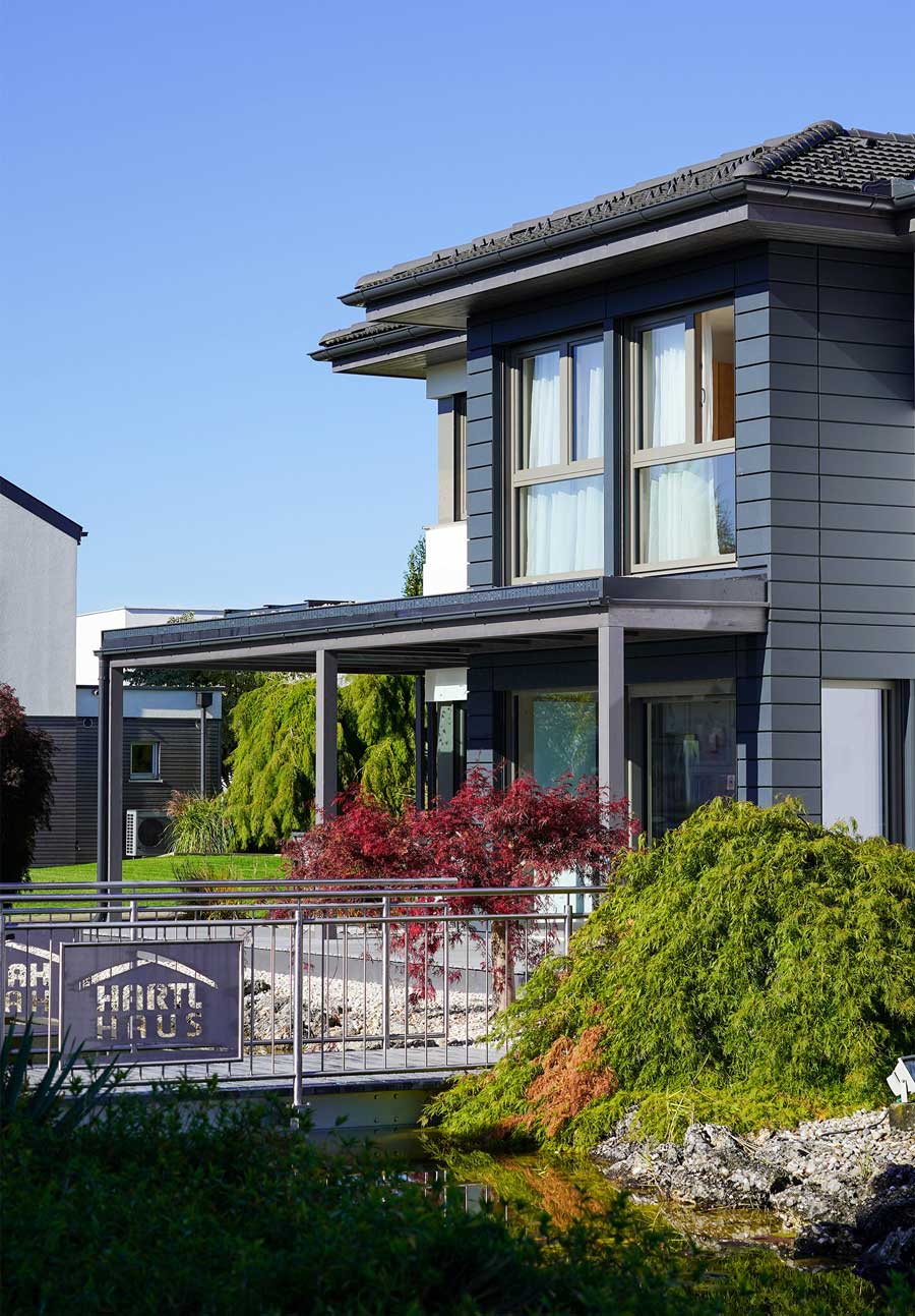 Modernes zweistöckiges Haus mit dunkelgrauer Fassade, großen Fenstern, gepflegtem Garten, kleinem Teich und Geländer mit der Aufschrift "Hartl Haus"